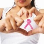 Woman in a white tank top forming a heart shape with hands, displaying a pink ribbon for Breast Cancer Awareness Month.