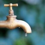 Brass faucet with dripping water, set against blurred green/blue foliage, highlighting plumbing services.