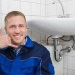 Smiling male plumber in blue and black uniform gestures to assist, near sink and tiled wall. B&L Plumbing services.