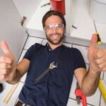 Smiling man with safety glasses lying on floor, tools scattered, showcasing DIY skills relevant to plumbing tasks.