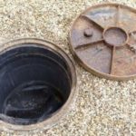 Open manhole surrounded by gravel and a rusty cover, illustrating urban plumbing maintenance issues.