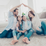 Family of four in a cozy living room, parents forming a roof with their hands over smiling children.