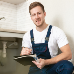 Smiling man in work attire with notepad at kitchen sink, representing B&L Plumbing's emergency service readiness.