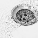 Close-up of a drain in water flow, highlighting its metallic texture and the importance of drain maintenance by B&L Plumbing.
