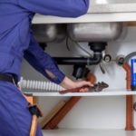 Technician in blue uniform repairing plumbing fixtures under a sink for B&L Plumbing drain cleaning services.