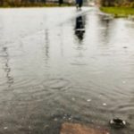 A person walking on a wet pathway with puddles, reflecting cloudy skies, related to plumbing service.