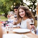 Group of people at a dining table outdoors, enjoying food and drinks, emphasizing a festive gathering during summer.