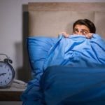 Young man in bed with blue bedding, looking fearful in a dim bedroom with a bedside clock, emphasizing plumbing concerns.