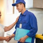 Delivery person in uniform shakes hands with a customer in a kitchen, holding a delivery folder for plumbing services.