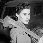 A woman with styled hair holds a large wrench over her shoulder next to an automobile, representing women in plumbing.
