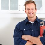 Smiling plumber in a blue shirt with a pipe wrench in a bright kitchen, showcasing B&L Plumbing's professional services.