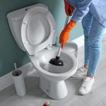 Person in orange gloves cleaning a toilet with a plunger in a bright bathroom, representing B&L Plumbing's maintenance tips.