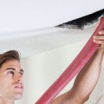Young man inspecting a damaged ceiling with red plumbing hose, representing B&L Plumbing's HVAC maintenance services.